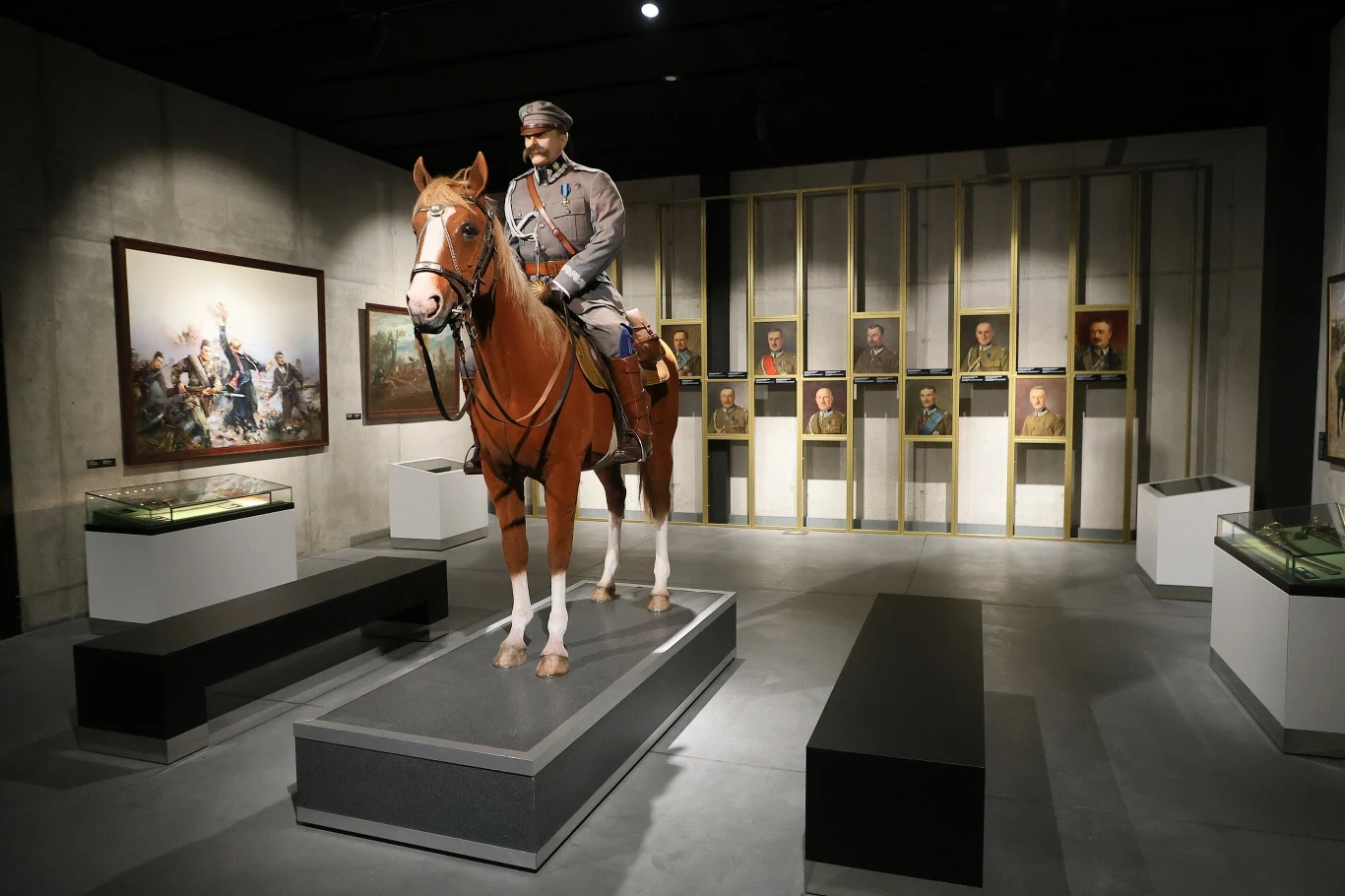 Manekin marszałka Józefa Piłsudskiego w mundurze wojskowym na koniu, ustawiony na podwyższeniu w muzeum, w tle galeria portretów historycznych postaci oraz obraz bitewny, całość w nowoczesnej aranżacji ekspozycyjnej.