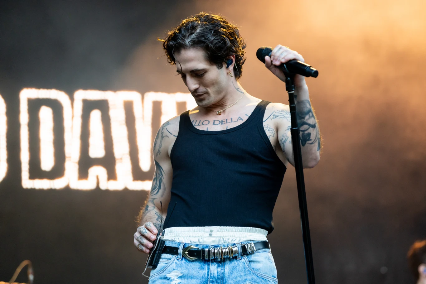 Damiano David rozwija swoją solową karierę poza Maneskin Młody mężczyzna w czarnej koszulce na ramiączkach i jasnych jeansach stoi na scenie z mikrofonem w dłoni, w tle rozświetlony napis tworzący fragment nazwy lub hasła, widoczne liczne tatuaże na rękach.