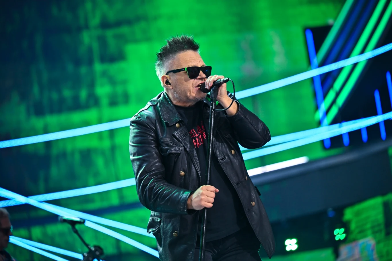 Muniek Staszczyk Wokalista w czarnej skórzanej kurtce i okularach przeciwsłonecznych śpiewa do mikrofonu na tle sceny koncertowej z dynamicznym, zielonym oświetleniem i neonowymi liniami świetlnymi.