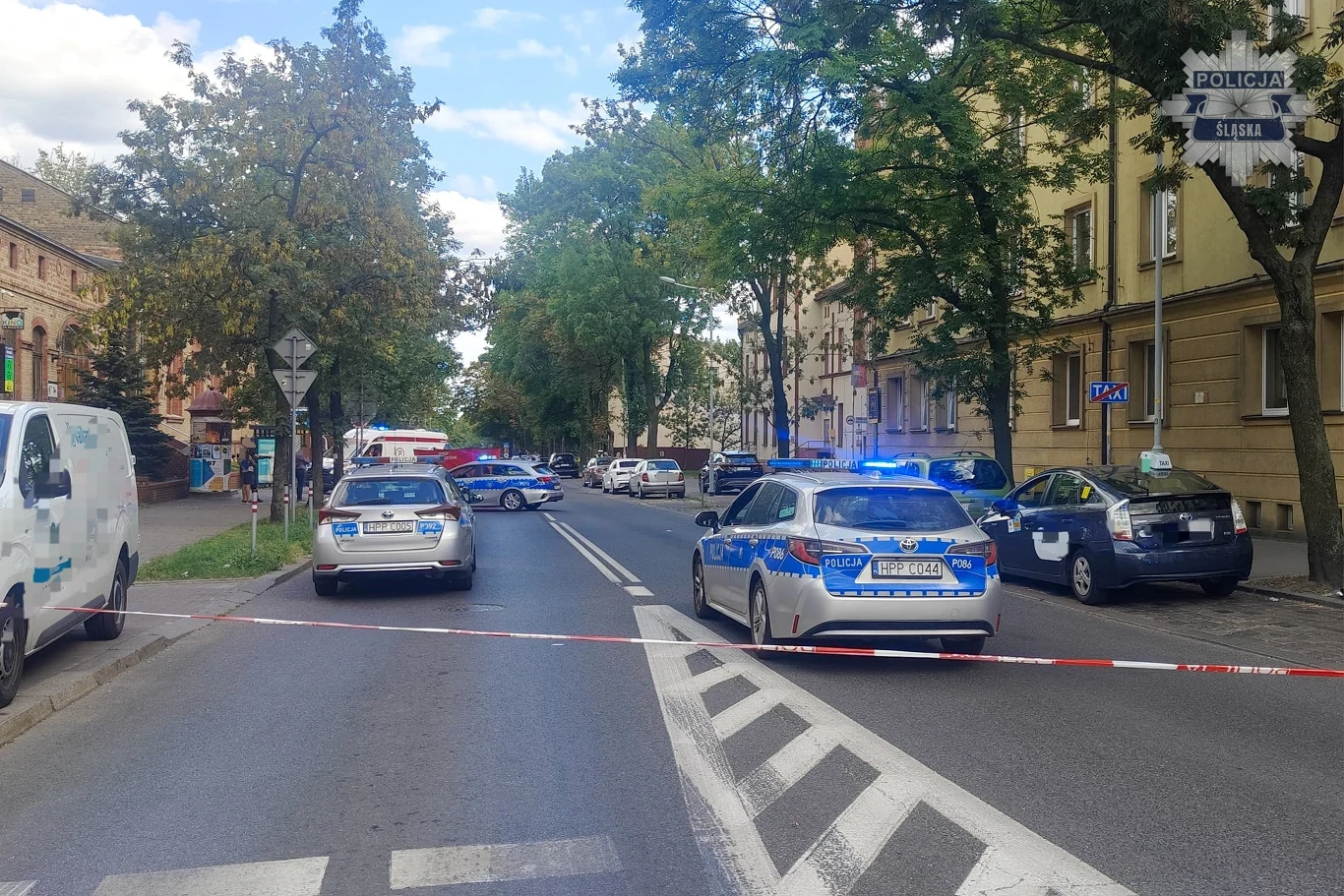 Atak nożownika w Chorzowie Kordon policyjny zabezpieczający fragment miejskiej ulicy, policyjne radiowozy z włączonymi sygnałami świetlnymi, taśma odgradzająca teren interwencji, w tle budynki, kilka innych pojazdów i drzewa.