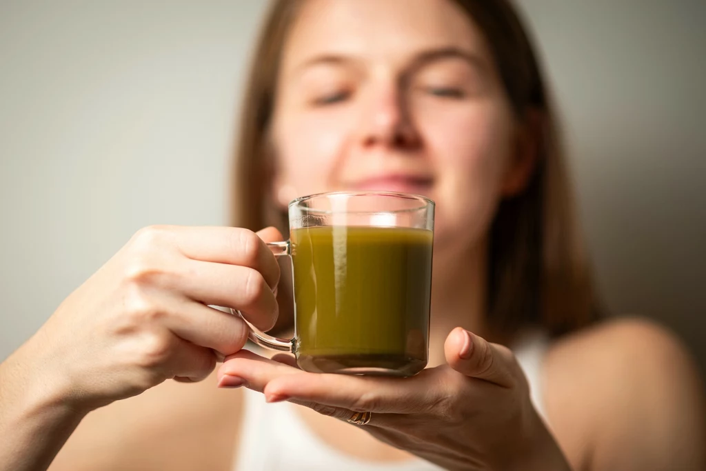 Wyciąg z kratomu może wyglądać tak Kobieta trzyma w ręku szklankę z zielonym napojem, skupiając się na jego zapachu lub smaku, delikatny uśmiech na twarzy, tło rozmyte.