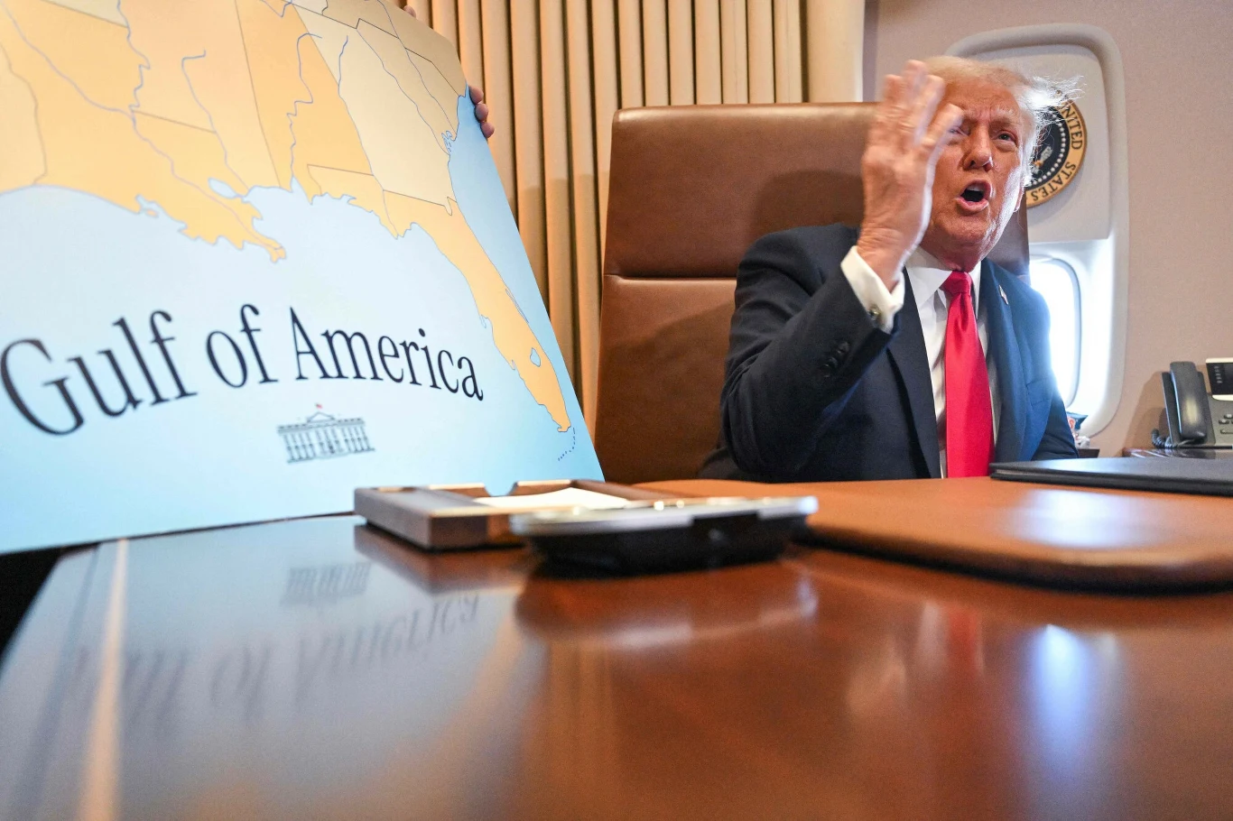 Mężczyzna w garniturze siedzący przy stole na pokładzie samolotu, energicznie gestykuluje ręką obok dużej tablicy z napisem 'Gulf of America' i zarysem południowej części Stanów Zjednoczonych.