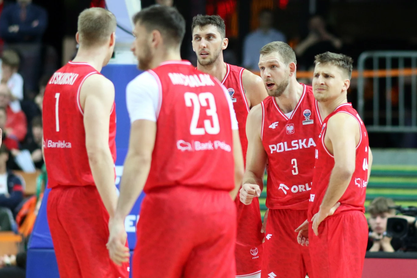 Grupa koszykarzy w czerwonych strojach z napisem Polska i numerami, stojących razem na parkiecie w trakcie meczu, skoncentrowanych na rozmowie lub ustaleniu dalszego przebiegu gry.
