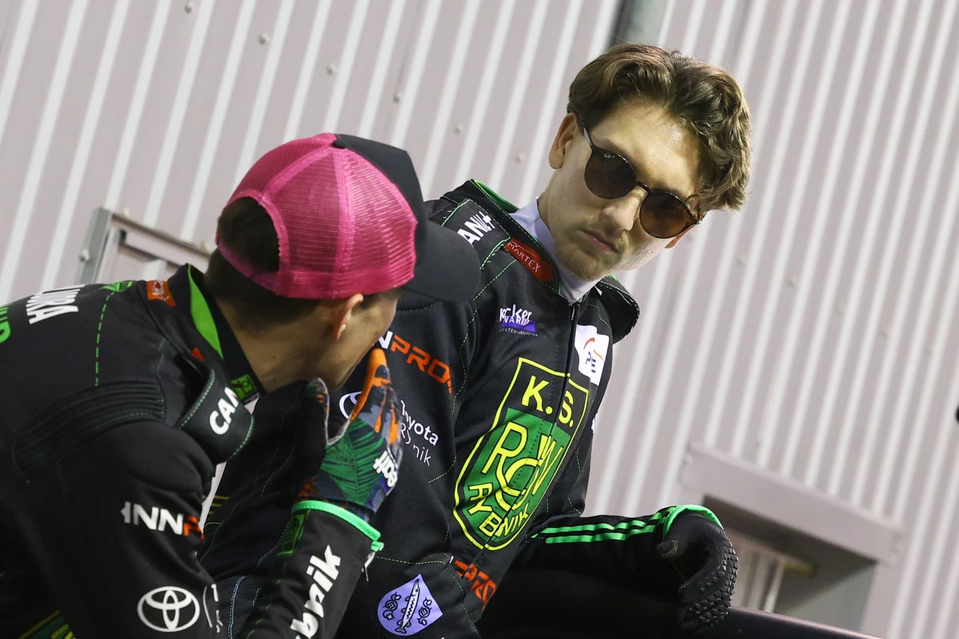 Kacper Pludra (w czapce) i Maksym Drabik Dwóch mężczyzn w kombinezonach żużlowych z logotypami sponsorów, jeden z nich nosi różowo-czarną czapkę, drugi okulary przeciwsłoneczne, obaj zwróceni ku sobie, na tle metalowej ściany.