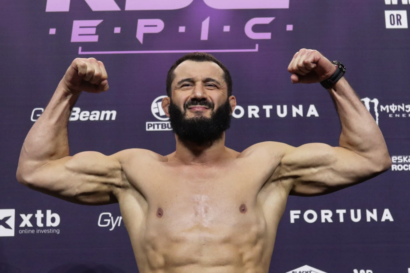 Atleta o umięśnionej sylwetce prezentuje napięte bicepsy na tle ściany z logotypami sponsorów i wydarzenia sportowego, wyraz twarzy pewny siebie.