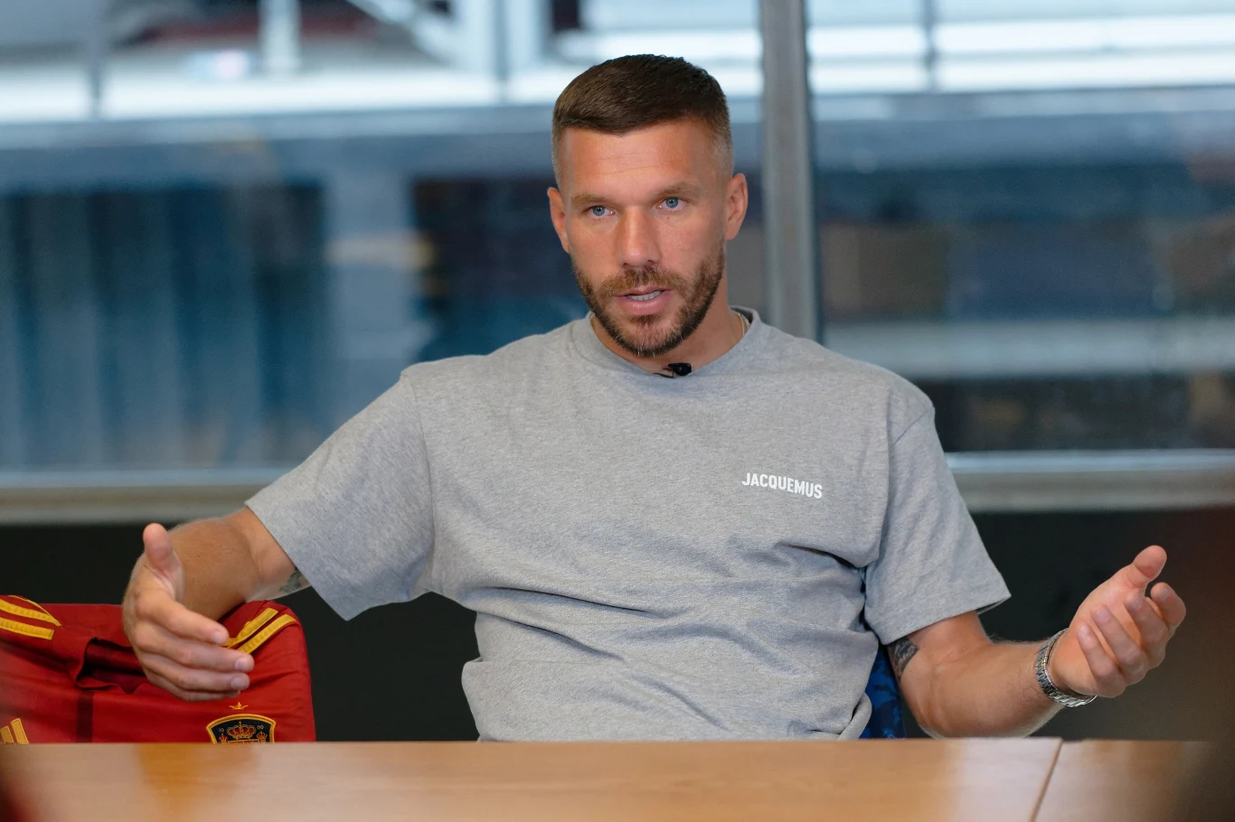 Lukas Podolski Mężczyzna z krótkimi włosami i zarostem siedzi przy stole w jasnoszarej koszulce, wykonując gesty dłońmi, tło stanowi duże okno wpuszczające rozproszone światło.
