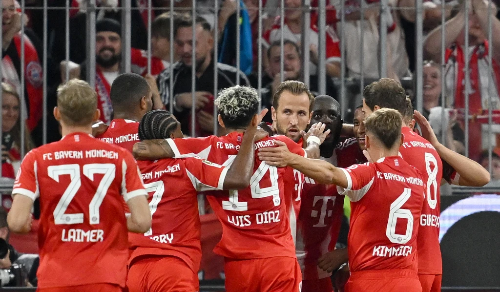 Piłkarze Bayernu Monachium świętują zdobycie bramki na boisku, obejmując się i ciesząc w grupie, na tle trybun wypełnionych kibicami.