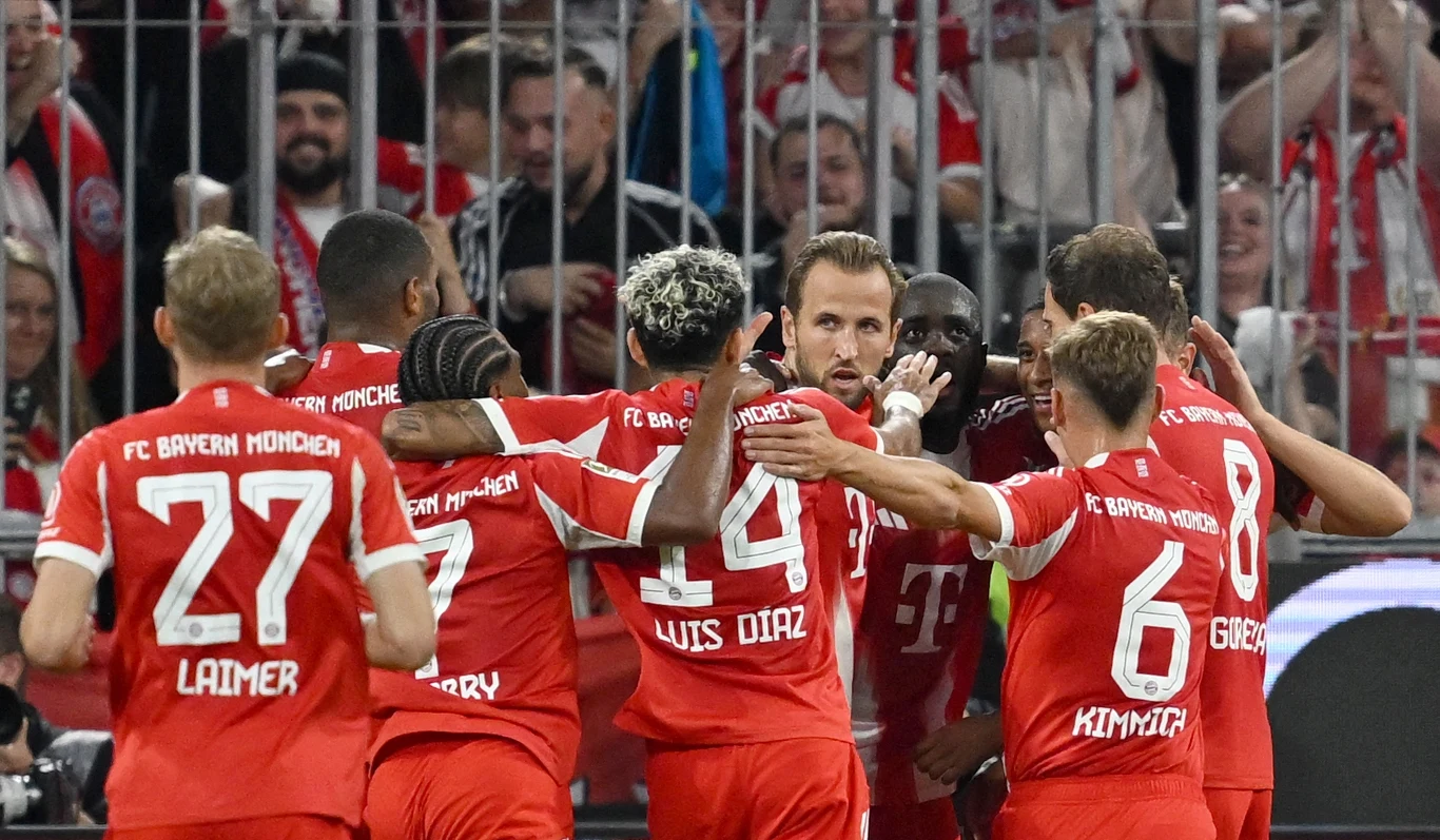 Piłkarze Bayernu Monachium świętują zdobycie bramki na boisku, obejmując się i ciesząc w grupie, na tle trybun wypełnionych kibicami.