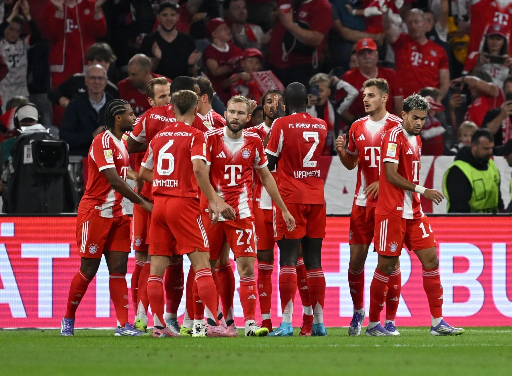 Piłkarze drużyny Bayern Monachium ubrani w czerwone stroje zgromadzeni razem na murawie w trakcie meczu, na trybunach widać kibiców ubranych w klubowe barwy.