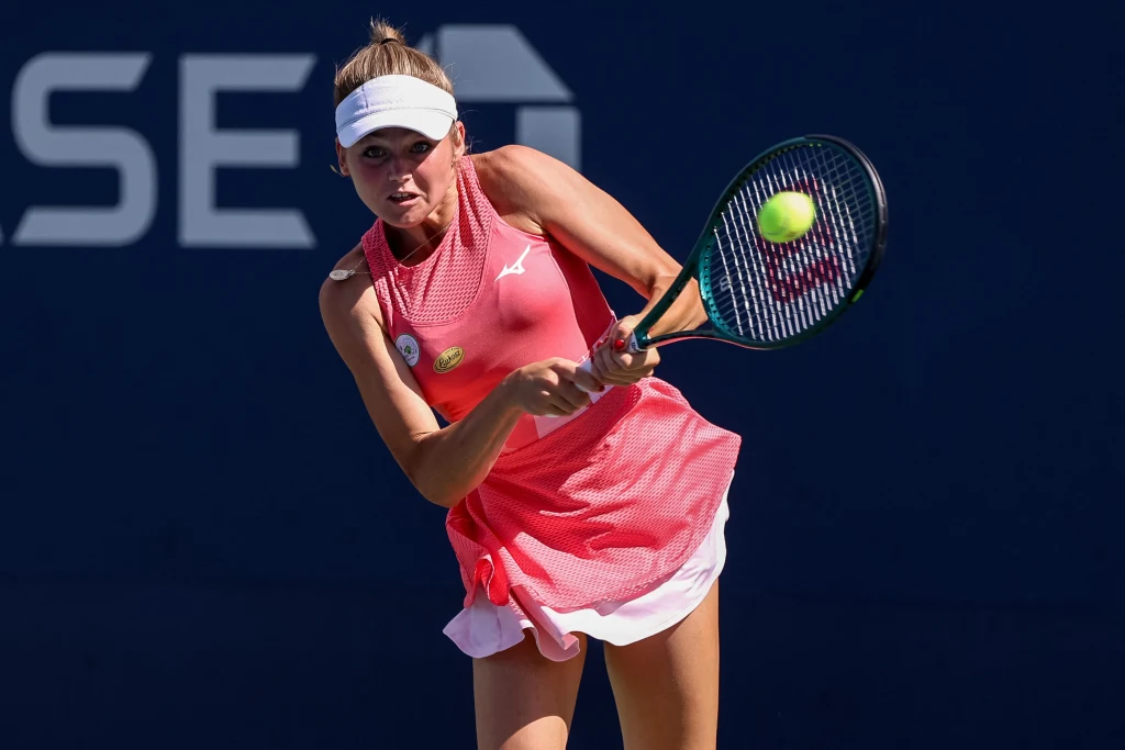 Tenisistka w czerwonej sukience wykonuje uderzenie piłki rakietą na korcie, skupienie w ruchu podkreślone przez sportowy strój i dynamiczną postawę, w tle fragment niebieskiego kortu.