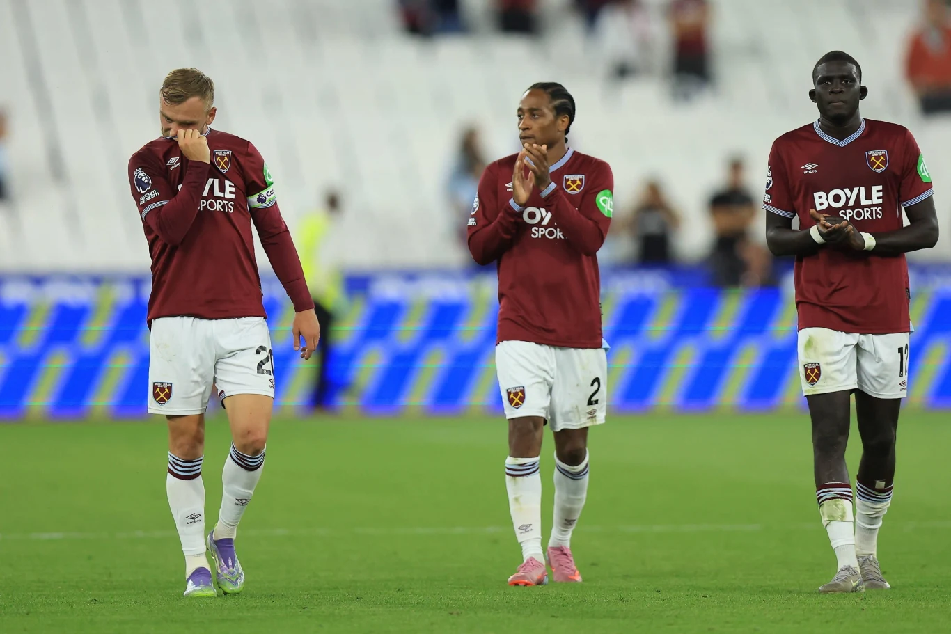 Trzech piłkarzy w bordowych koszulkach West Ham United idzie razem po murawie stadionu, wykazując emocje po zakończonym meczu, na pierwszym planie zawodnik ociera twarz dłonią.