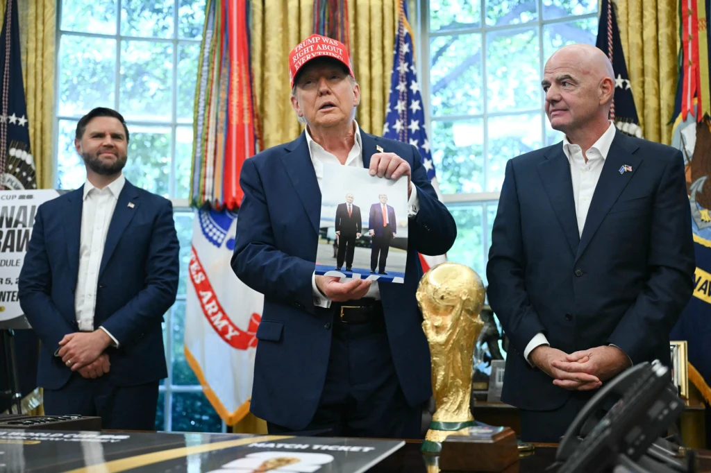 Prezydent USA Donald Trump podczas spotkania z szefem FIFA Giannim Infantino nagle pokazał swoje zdjęcie z Władimirem Putinem Trzech mężczyzn stoi w gabinecie o wysokiej randze, na pierwszym planie jeden z nich trzyma w rękach fotografię z innym mężczyzną, obok znajduje się trofeum w kształcie Złotej Piłki. W tle widoczne są flagi i bogato udekorowane wnętrze.
