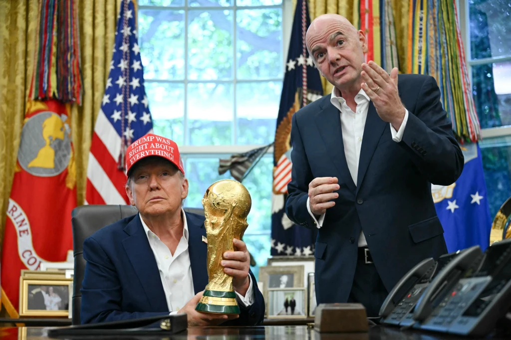 Donald Trump i Gianni Infantino Dwóch mężczyzn w garniturach stoi w gabinecie z licznymi flagami w tle, jeden z nich trzyma trofeum Mistrzostw Świata w piłce nożnej, drugi gestykuluje podczas rozmowy.