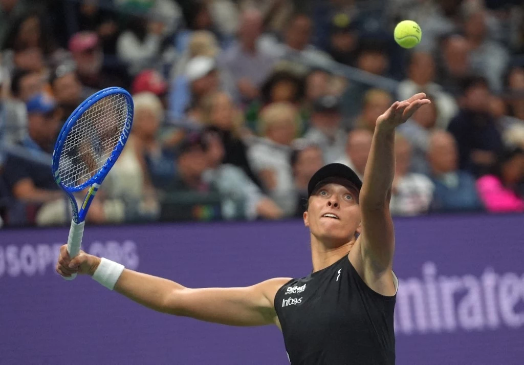 Iga Świątek jest jedną z faworytek US Open 2025 Tenisistka w czarnej koszulce podczas wykonywania serwisu na korcie tenisowym, z widoczną piłką w powietrzu, w tle rozmyta publiczność kibicująca na trybunach.