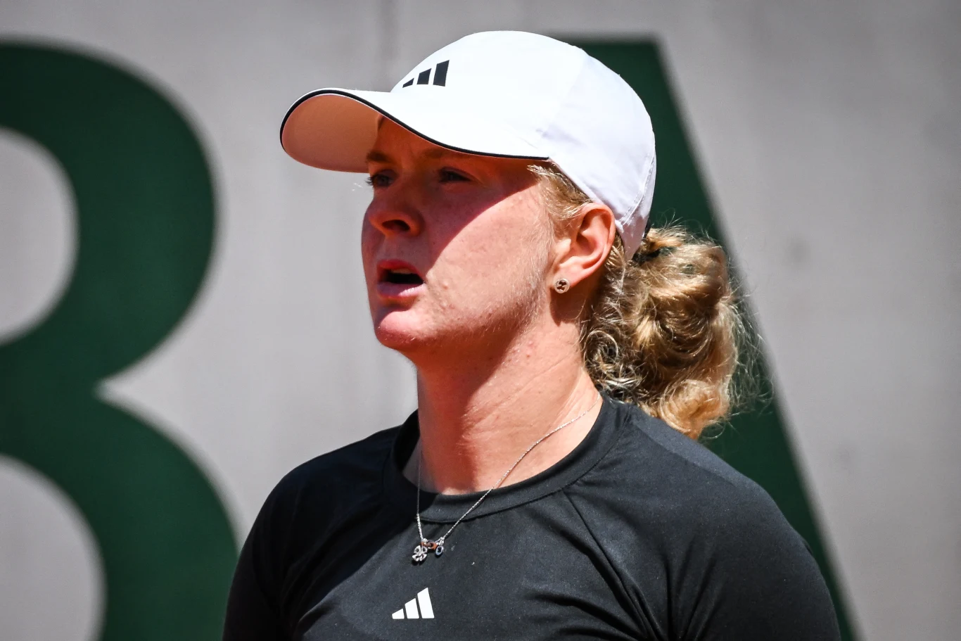 Francesca Jones Sportowiec w czapce z daszkiem i ciemnej koszulce, skupiona twarz, tło rozmyte z elementami zielonego koloru oraz cyfrą znajdującą się częściowo w kadrze.