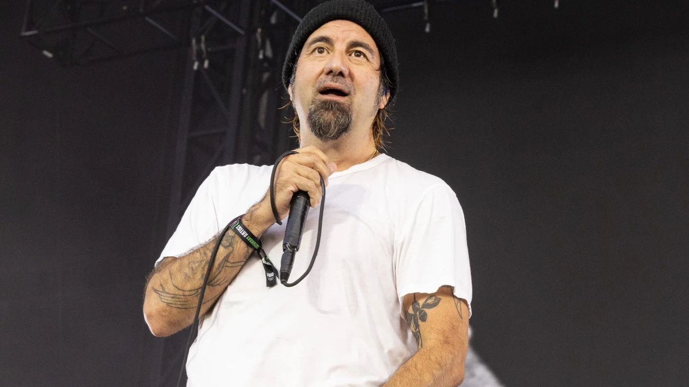 Chino Moreno (Deftones) Mężczyzna w białej koszulce i czarnej czapce z mikrofonem w ręce, z widocznymi tatuażami na przedramieniu, stoi na scenie podczas występu lub koncertu, jasne światło podkreśla jego sylwetkę na ciemnym tle.