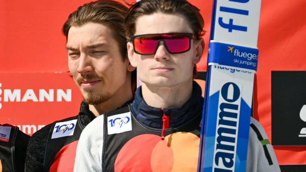 Johann Andre Forfang, Marius Lindvik Dwóch młodych mężczyzn w sportowych strojach narciarskich, jeden z nich trzyma narty, a drugi stoi obok; obaj mają przypięte numery startowe i znajdują się na tle reklam sponsorskich.