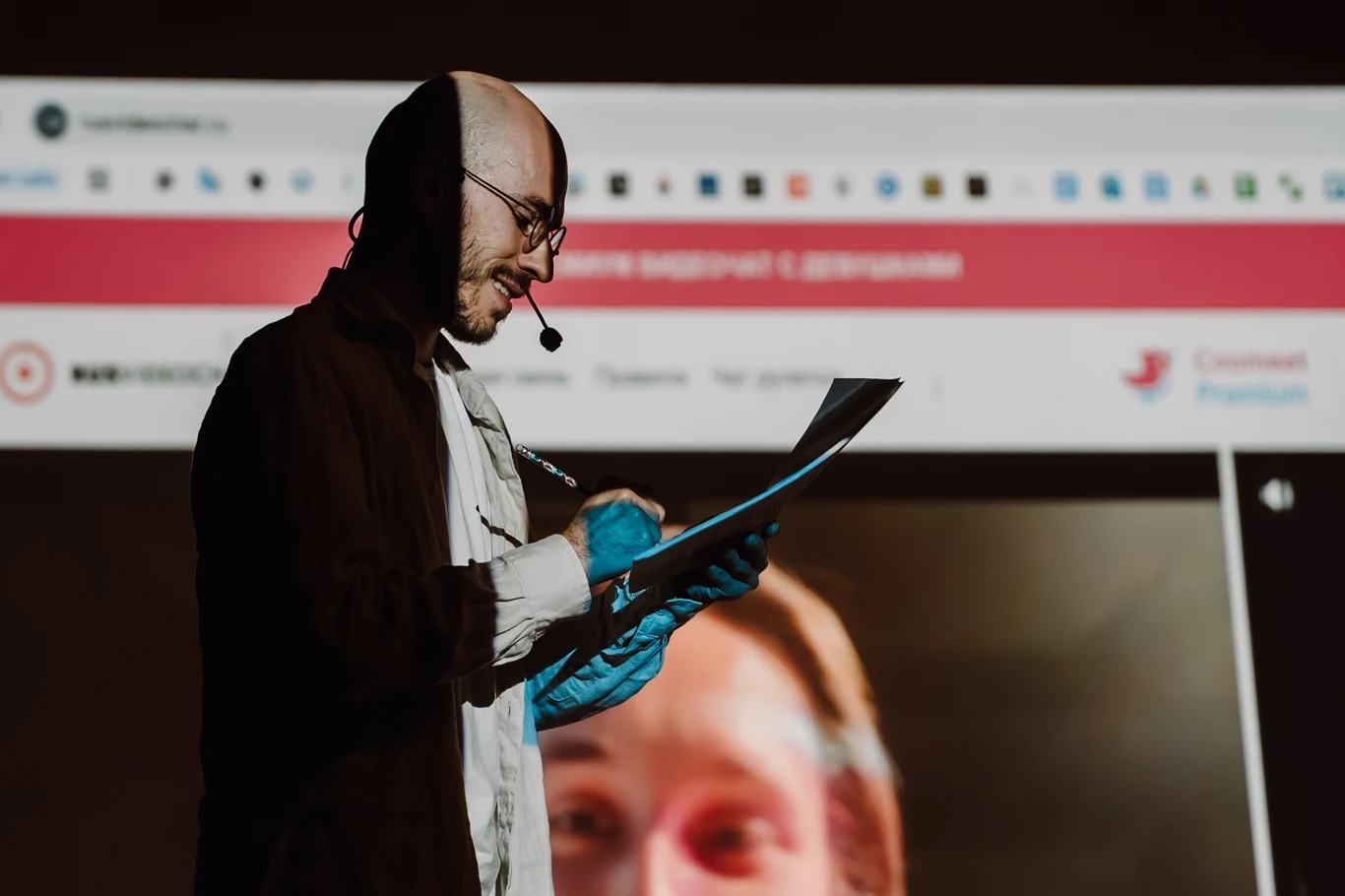 Mężczyzna w słuchawkach z mikrofonem i niebieskimi rękawiczkami na dłoniach pisze coś na kartce, stojąc przed dużym ekranem z obrazem twarzy kobiety i stroną internetową w tle.