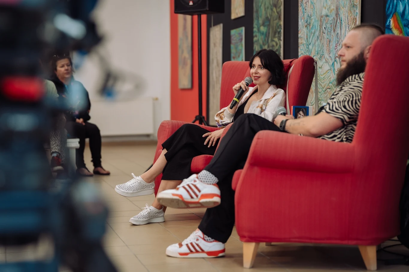 Dwoje osób siedzących na czerwonych fotelach podczas rozmowy z mikrofonem w ręku, w tle widoczne obrazy na ścianie oraz niewyraźna postać obserwująca wydarzenie.