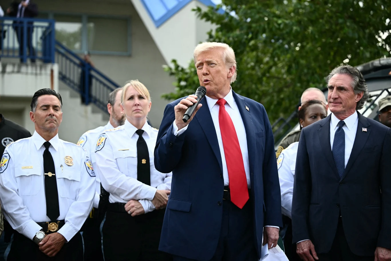 Prezydent USA Donald Trump Grupa osób w oficjalnych mundurach i garniturach zgromadzona na zewnątrz, centralnie mężczyzna w granatowym garniturze z czerwonym krawatem mówi do mikrofonu, obok niego stoją funkcjonariusze oraz inny mężczyzna w garniturze