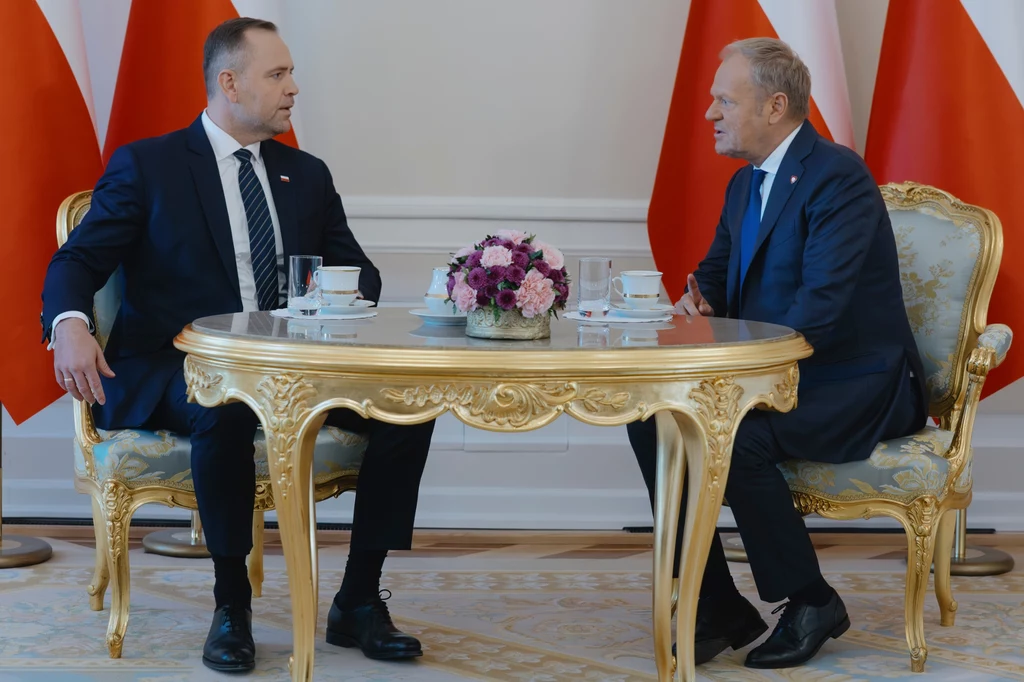Karol Nawrocki i Donald Tusk Dwóch mężczyzn siedzi naprzeciwko siebie przy ozdobnym, złotym stole, na którym stoi bukiet kwiatów i szklanki wody, w tle widoczne polskie flagi.