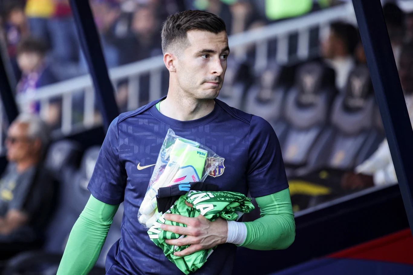 Inaki Pena Młody mężczyzna w stroju sportowym trzyma w rękach odzież i akcesoria piłkarskie, zasiada na stadionowej ławce rezerwowych, w tle widoczni są kibice i elementy stadionu.