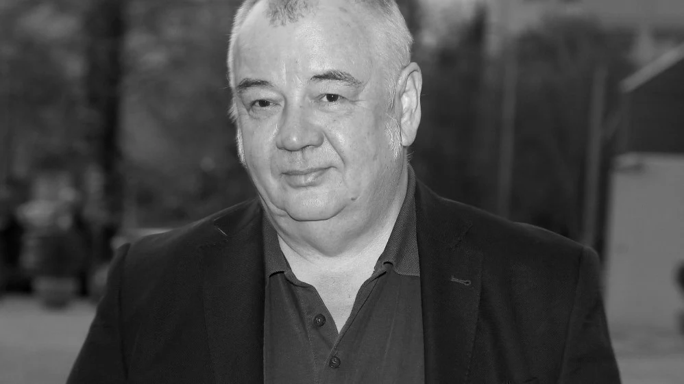 Stanisław Soyka Starszy mężczyzna o siwych włosach w ciemnej marynarce, z łagodnym wyrazem twarzy, stojący na zewnątrz w rozmytym, miejskim otoczeniu.