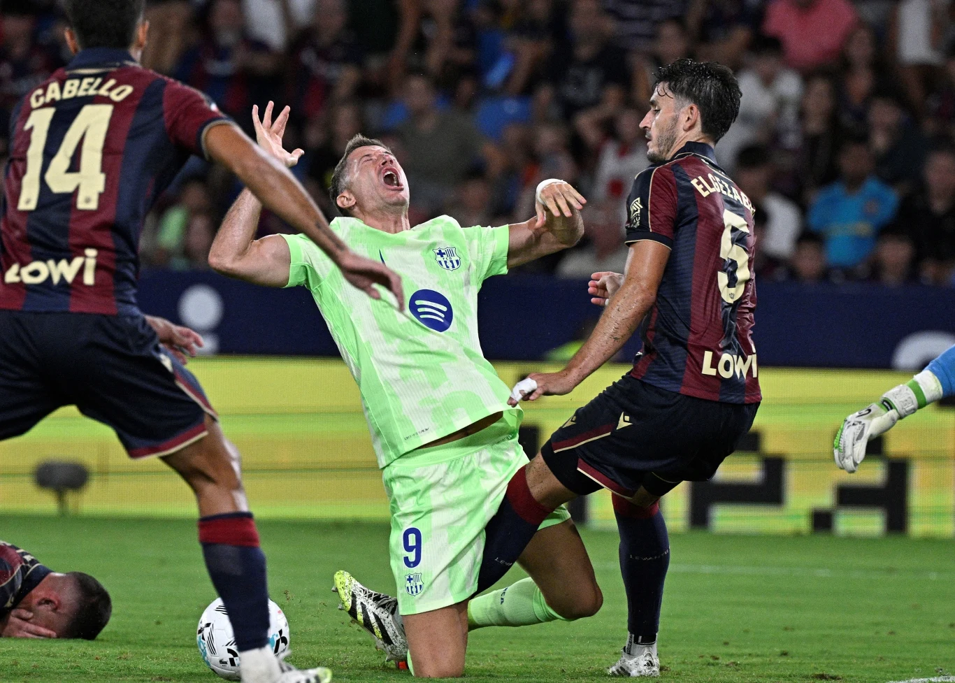 Zawodnik w jasnozielonym stroju pada na murawę z widocznym grymasem bólu, podczas gdy piłkarz w ciemnym stroju z numerem 5 wykonuje wślizg, a na drugim planie znajdują się kolejni zawodnicy i kibice.