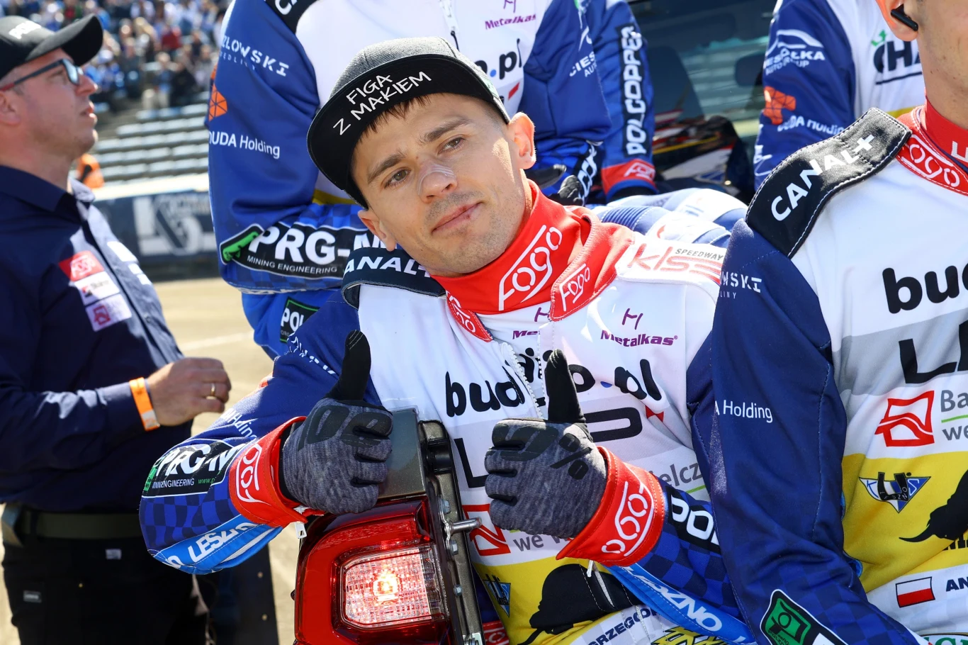 Mężczyzna w stroju żużlowym trzyma kciuki w górę, uśmiechnięty, otoczony innymi zawodnikami podczas zawodów sportowych na torze żużlowym, na czapce napis 'FIRGA Z MAKIEM'.