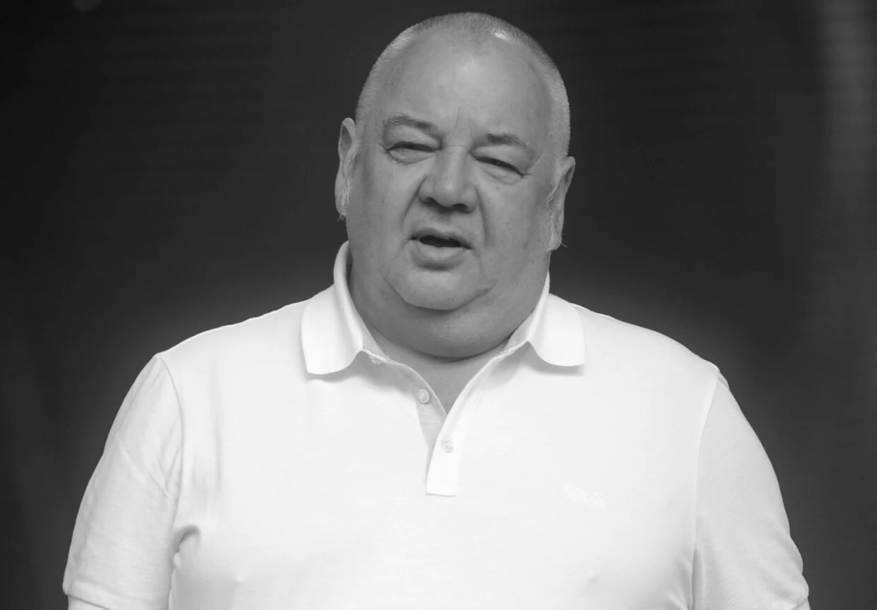 Stanisław Soyka zmarł w wieku 66 lat Mężczyzna w średnim wieku o pełnej sylwetce, ubrany w białą koszulkę polo, na tle ciemnego, rozmytego tła, patrzy w stronę obiektywu z lekko otwartymi ustami.