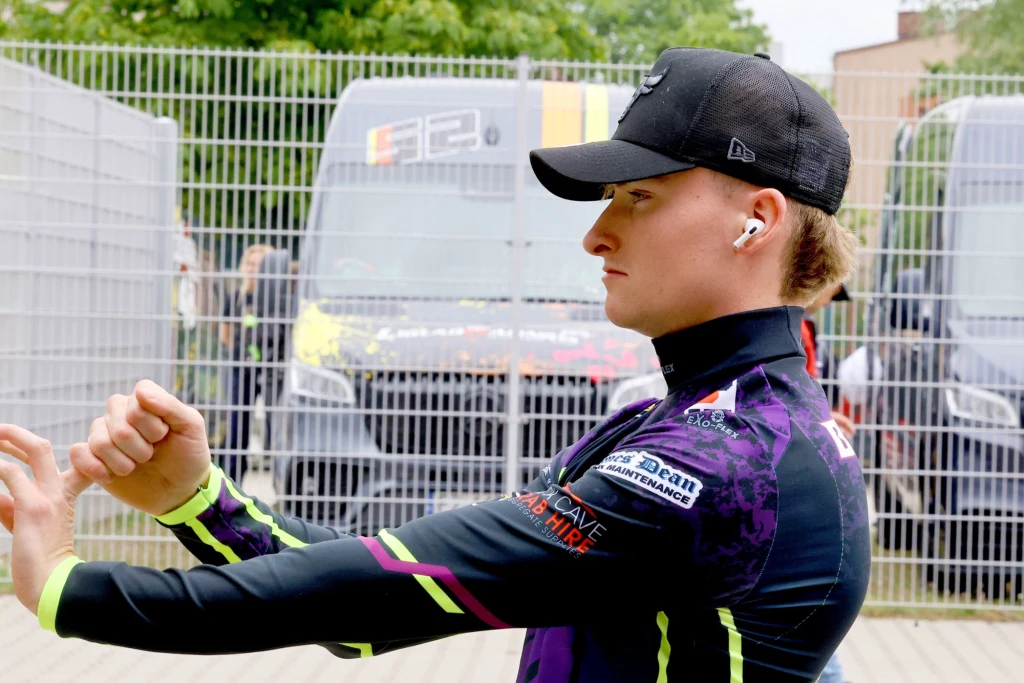 Młody mężczyzna ubrany w sportową odzież oraz czapkę z daszkiem stoi przodem w półprofilu, rozciąga ręce, w tle widoczne są samochody dostawcze oraz metalowe ogrodzenie.