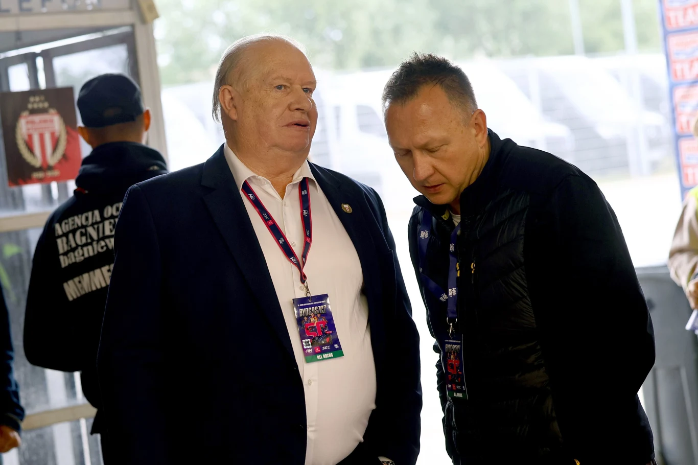 Prezes Polonii Jerzy Kanclerz wraz z Jackiem Frątczakiem. Dwóch mężczyzn w eleganckich ubraniach rozmawia ze sobą w zamkniętym pomieszczeniu, obaj mają na szyjach identyfikatory, w tle widoczny mężczyzna w kurtce z napisem agencja ochrony oraz wejście na stadion lub do hali sportowej.