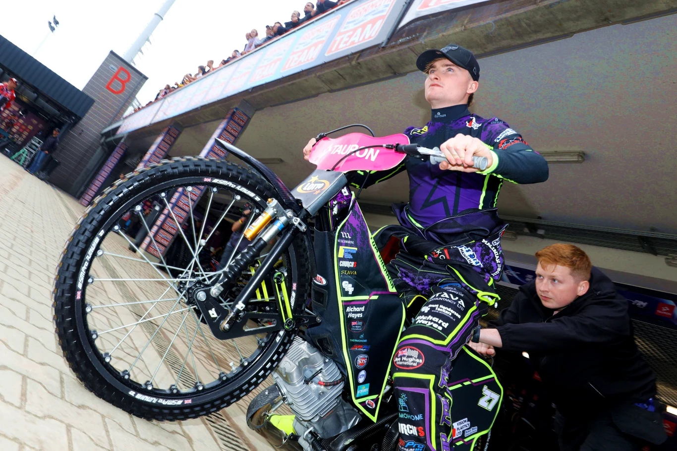 Tom Brennan Zawodnik żużlowy w pełnym stroju sportowym siedzi na motocyklu żużlowym, obok stoi druga osoba pomagająca wypchnąć motocykl z boksu. Na trybunach widoczna grupa kibiców obserwujących zawodników.