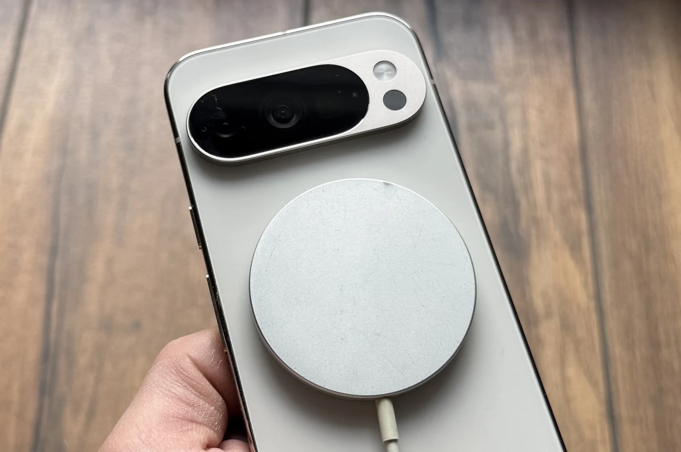 Smartfon trzymany w dłoni, do którego tyłu przyczepiona jest magnetyczna ładowarka bezprzewodowa, widoczna blisko modułu aparatu. Tło stanowi drewniana powierzchnia.
