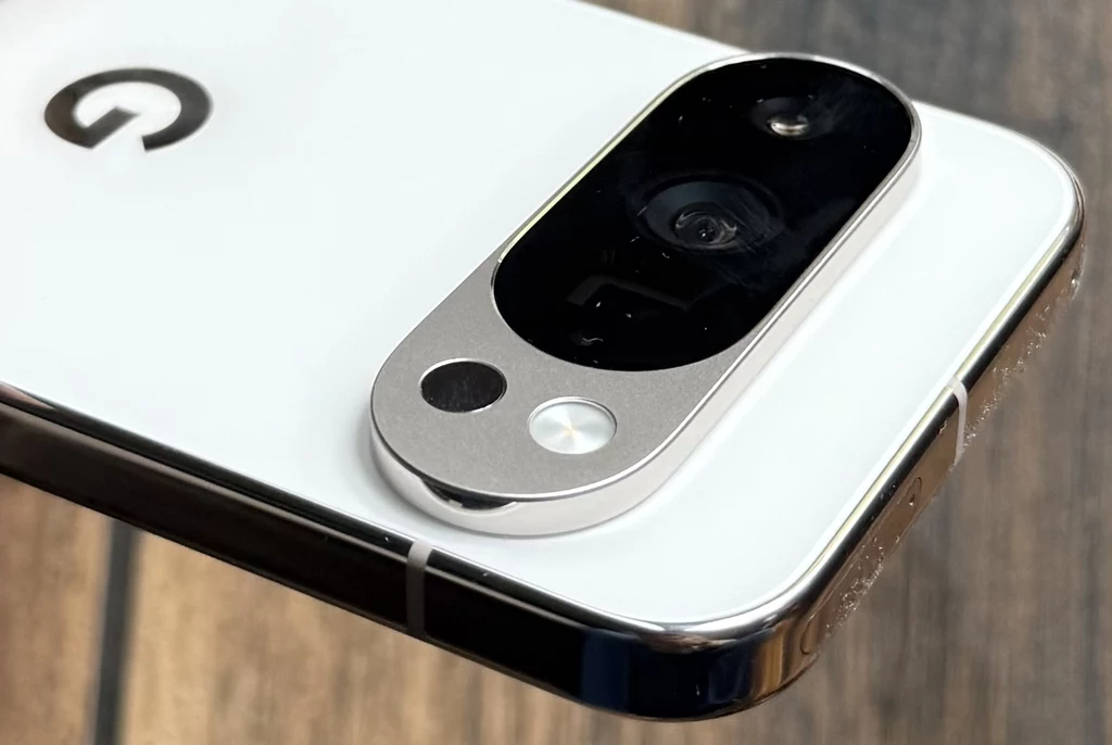 Google Pixel 10 Pro to smartfon, który jest ładnie zaprojektowany i przykuwa wzrok. Moduł aparatu fotograficznego na białej obudowie smartfona z wyraźnym logo w górnej części oraz detalami metalu i szkła, całość na tle drewnianego podłoża.