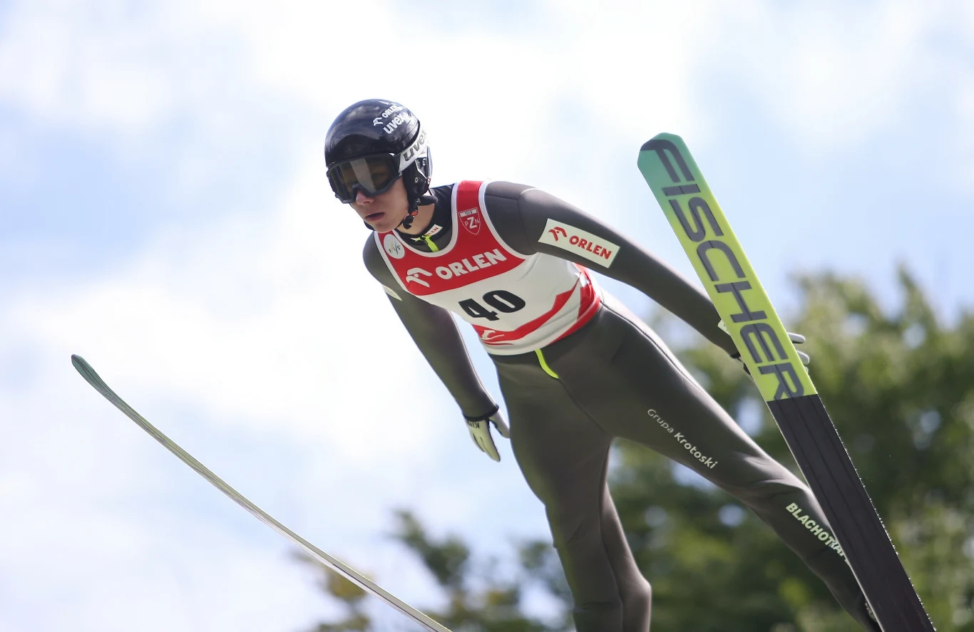 Zawodnik podczas skoku narciarskiego w locie, ubrany w kombinezon oraz kask z goglami, z numerem startowym na piersi, w tle niebo i rozmyte drzewa.