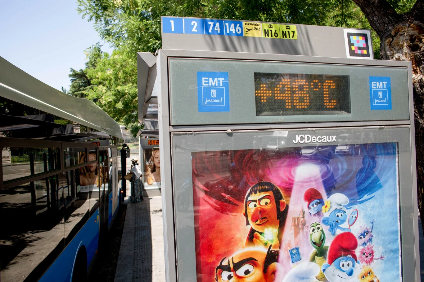 Przystanek autobusowy z elektronicznym wyświetlaczem pokazującym temperaturę 48°C, obok autobus miejski oraz plakat filmowy z postaciami animowanymi w centralnej części konstrukcji.