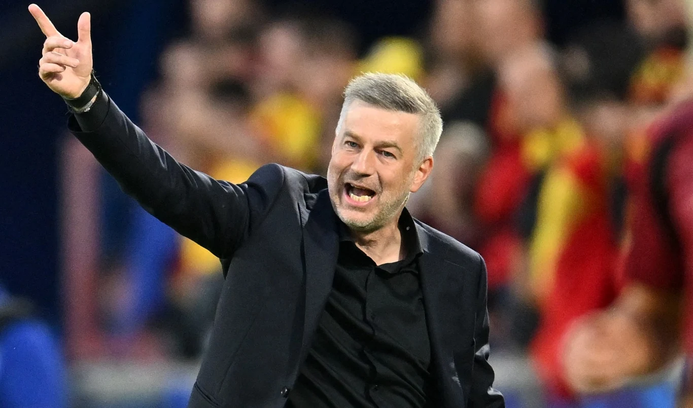 Mężczyzna w ciemnej marynarce energicznie gestykuluje, unosi ramię i wskazuje palcem, za nim znajdują się rozmyte sylwetki ludzi w tle.