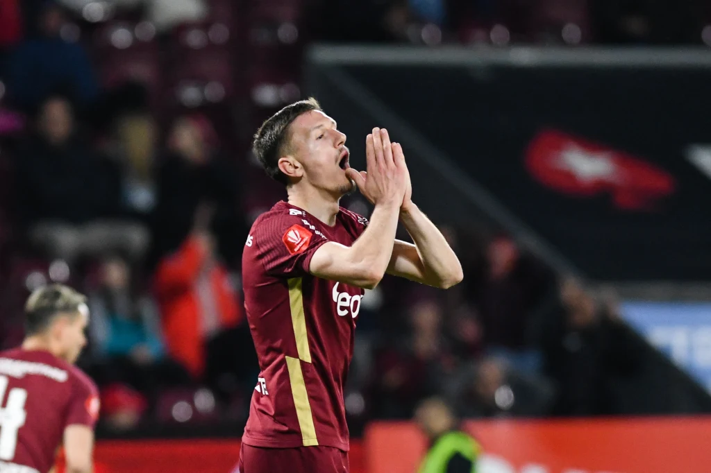 Piłkarz ubrany w bordowy strój sportowy stoi na boisku z dłońmi złożonymi przy twarzy w geście rozczarowania lub modlitwy, w tle widoczni inni zawodnicy oraz rozmyty tłum na trybunach.