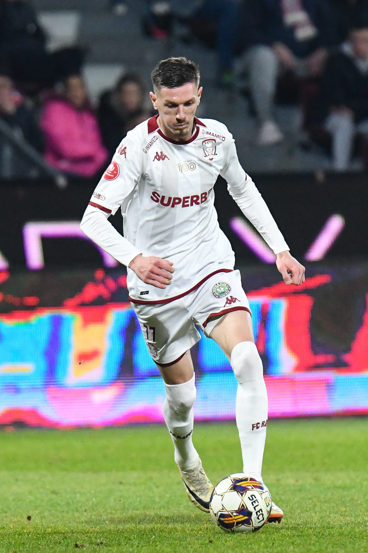 Piłkarz w białym stroju z logo klubu kontroluje piłkę nożną podczas meczu na zielonej murawie stadionu, w tle widownia i kolorowe światła.