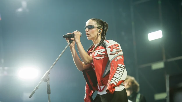 Kobieta w sportowej kurtce z napisem Yamaha, nosząca okulary przeciwsłoneczne, śpiewa do mikrofonu na scenie podczas koncertu, w tle widoczny gitarzysta i światła sceniczne.