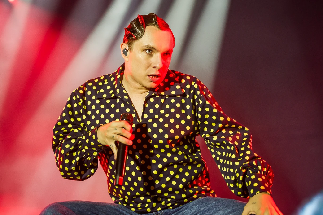 Ralph Kamiński podczas finału Męskiego Grania 2025 Artysta na scenie w kolorowej koszuli w grochy i charakterystycznej fryzurze trzymający mikrofon, oświetlony światłami koncertowymi w czerwonej tonacji