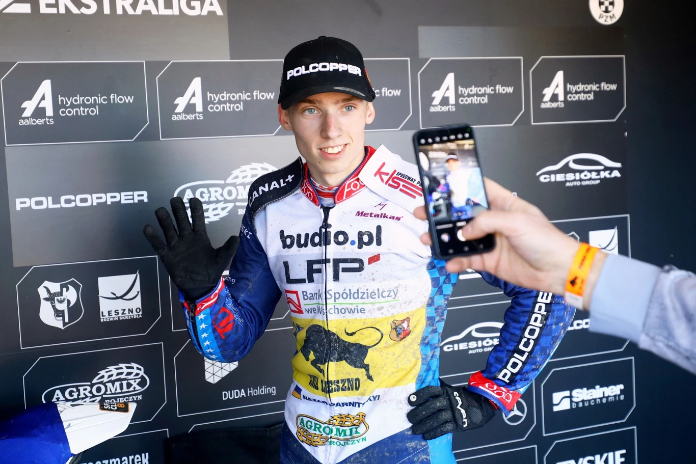 Zawodnik żużlowy w stroju sportowym pozuje do zdjęcia, trzyma rękę w geście powitania, obok osoba robi mu zdjęcie telefonem, w tle widoczne logotypy sponsorów i organizatorów wydarzenia.