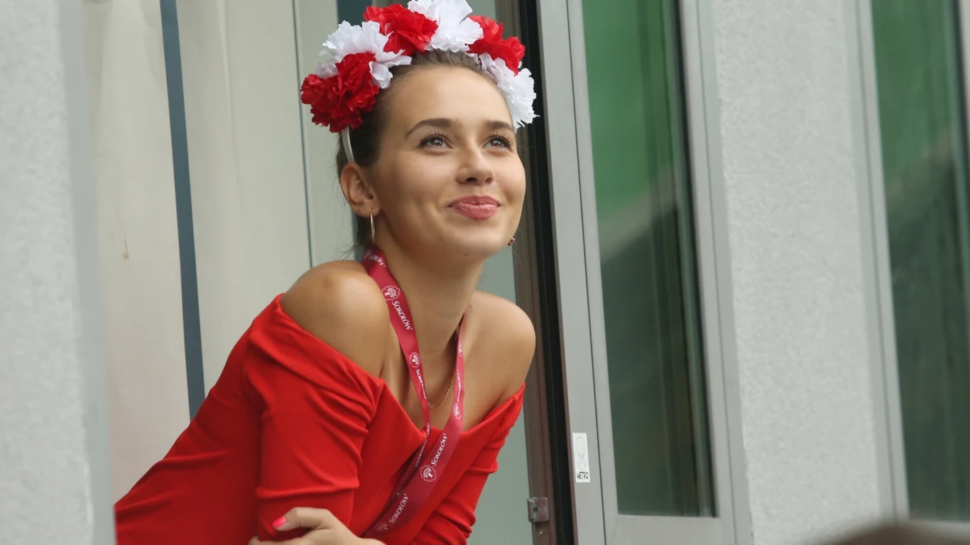 Młoda kobieta w czerwonej bluzce i ozdobnej opasce z czerwonych oraz białych kwiatów na głowie spogląda z uśmiechem przez okno, mając przewieszoną przez szyję smycz z identyfikatorem.