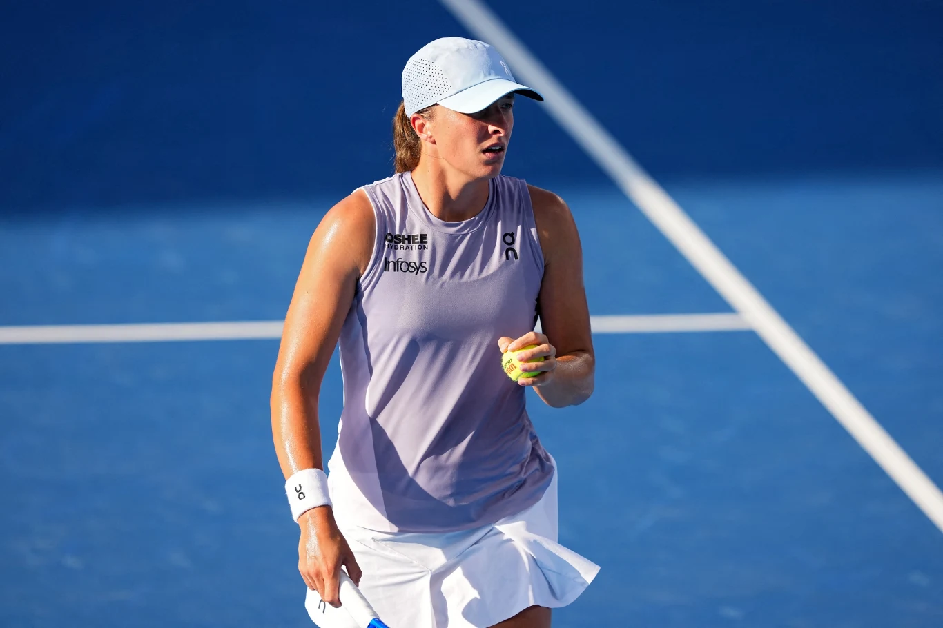 Tenisistka ubrana w sportowy strój, trzymająca w ręku piłkę tenisową, stoi na niebieskim korcie podczas meczu; intensywne światło słoneczne oraz czapka z daszkiem chroniąca przed słońcem.