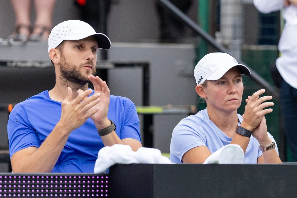 Maciej Ryszczuk w towarzystwie Darii Abramowicz, odpowiadającej za przygotowanie mentalne Igi Świątek Dwie osoby w sportowych ubraniach i białych czapkach siedzą obok siebie, klaszcząc na trybunach, w tle widać fragmenty trybun oraz inne osoby.