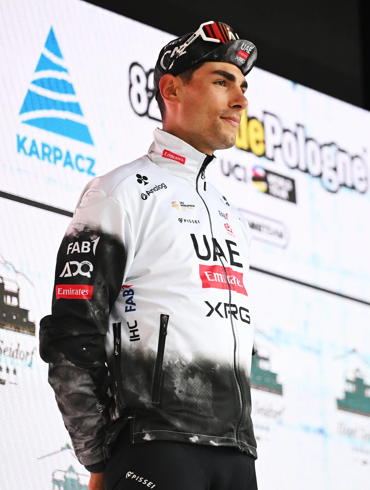 Zawodnik UAE Team Emirates w sportowej odzieży kolarskiej stoi na tle planszy z logo wyścigu Tour de Pologne oraz logotypami sponsorów.