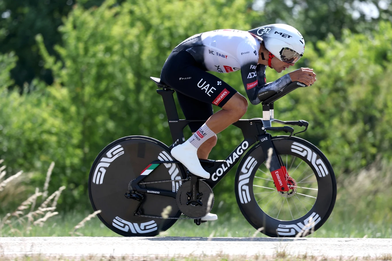 Kolarz jadący na rowerze czasowym w profesjonalnym stroju aerodynamicznym podczas wyścigu, na tle zieleni i drzew.