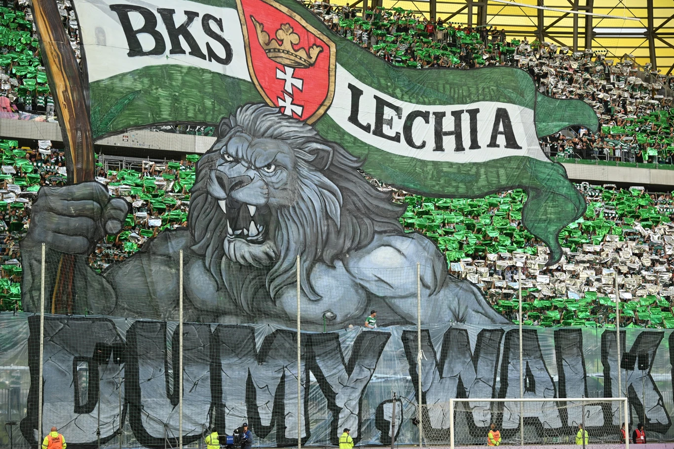 Ogromny transparent z wizerunkiem lwa trzymającego flagę z napisem BKS Lechia oraz herbem klubu na tle wypełnionego stadionu, kibice w zielono-białych barwach podkreślają oprawę dopingu.