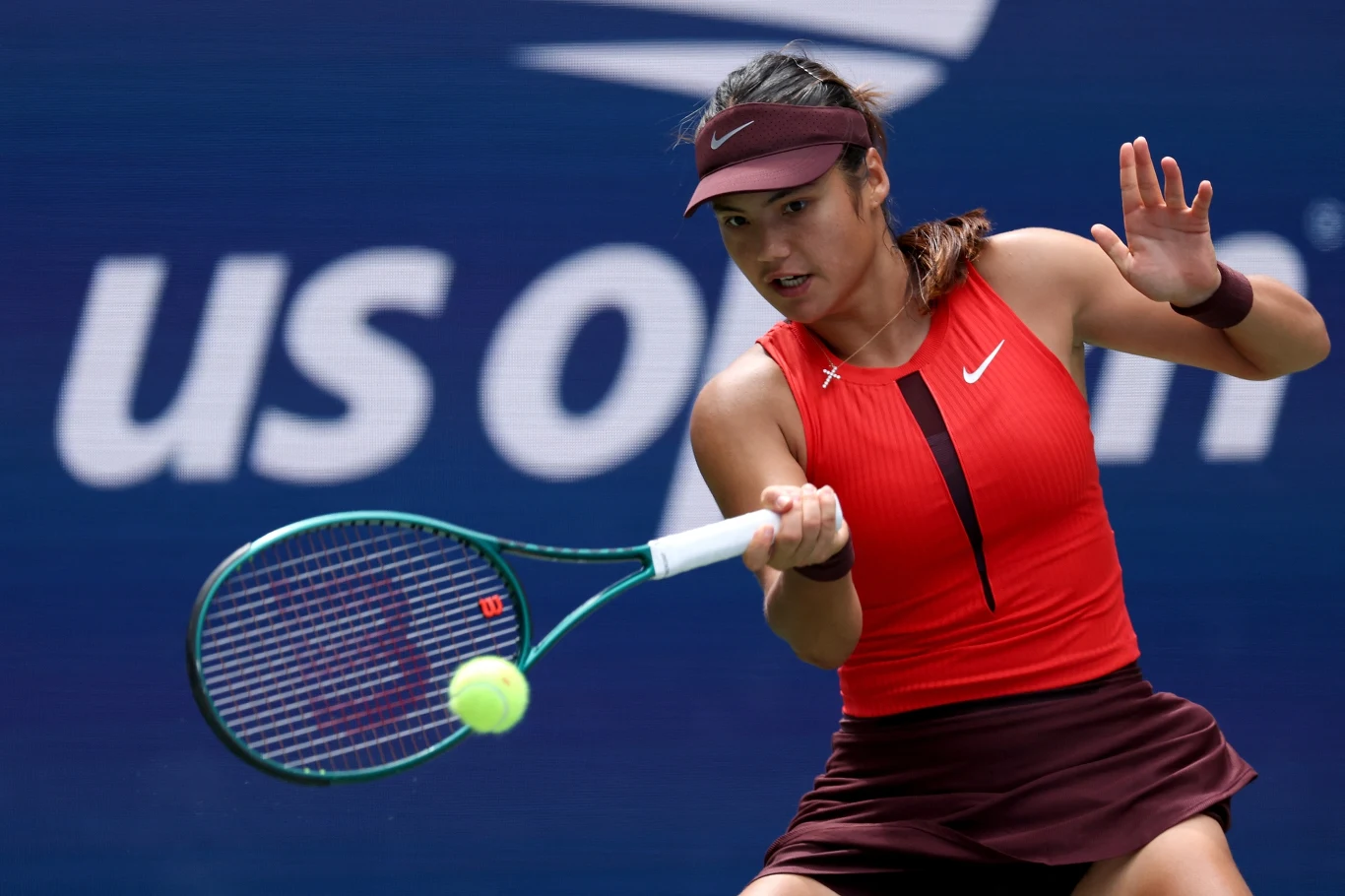 Emma Raducanu Tenisistka ubrana w czerwoną koszulkę i bordową spódniczkę odbija piłkę podczas meczu na tle napisu US Open.